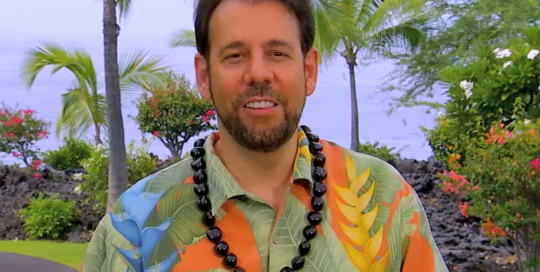 Perillo Tours Hawaii Commercial thumbnail features Steve Perillo wearing a bright Hawaiian print shirt and a traditional black Kukui nut lei against a backdrop of beautiful Hawaiian trees and bushes (video production by Merging Media).