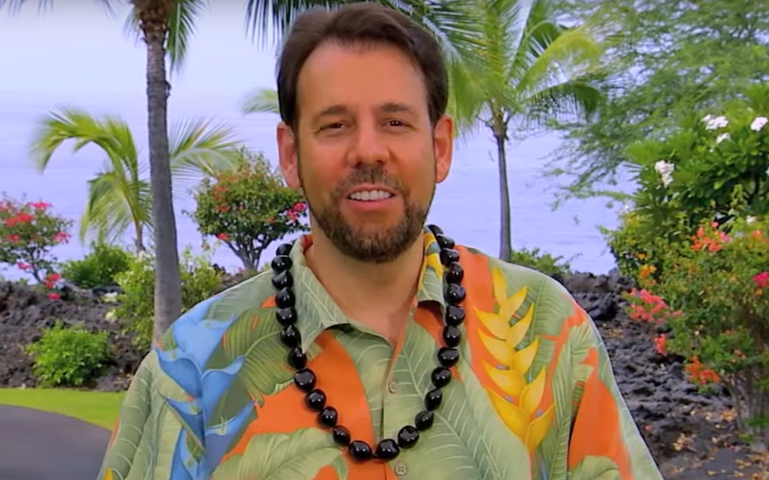 Perillo Tours Hawaii Commercial thumbnail features Steve Perillo wearing a bright Hawaiian print shirt and a traditional black Kukui nut lei against a backdrop of beautiful Hawaiian trees and bushes (video production by Merging Media).