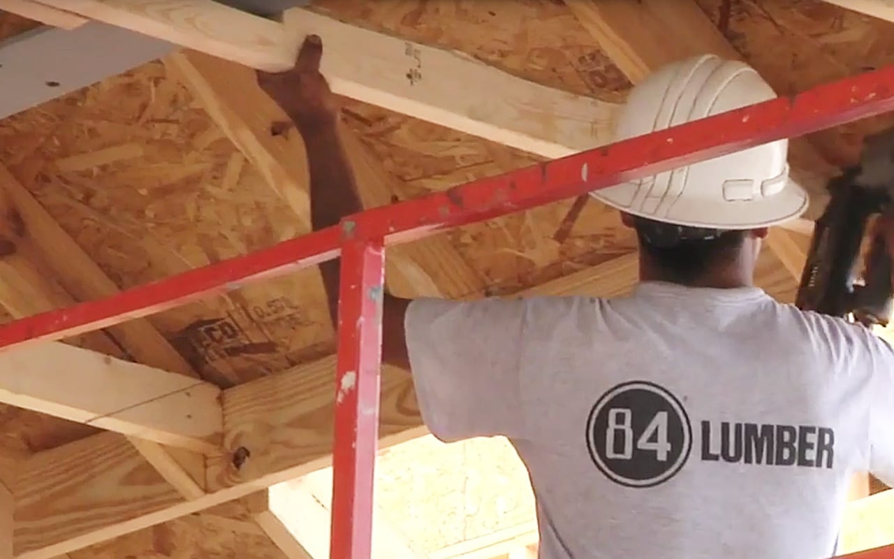 84 Lumber Construction Services video thumbnail features a man in a white tee-shirt and a white hard had with his back to the camera as he installs a roof joist, company logo on his shirt reads; 84 Lumber (video production by Merging Media).
