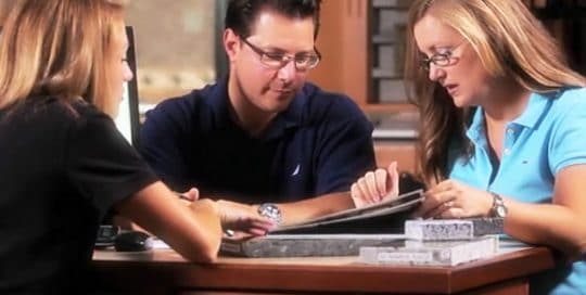 84 Lumber Kitchen How-To video thumbnail features a husband and a wife collaborating at a desk over samples of kitchen countertop materials while an employee from 84 Lumber looks on to give professional advice (video production by Merging Media).