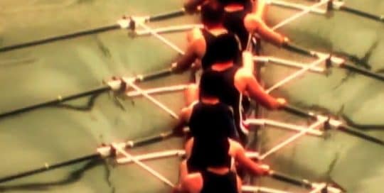 Birdseye view of a mens rowing team, wearing black tank top uniforms, in an eight person shell in Bobrick 2006 Vision Conference commercial
