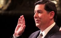 Mayor Peduto stands in his suit and tie at his inauguration with right hand in the air to be sorwn in as the mayor of Pittsburgh, Pennsylvania as recorded in Mayor Peduto Inauguration video (video production by Merging Media).