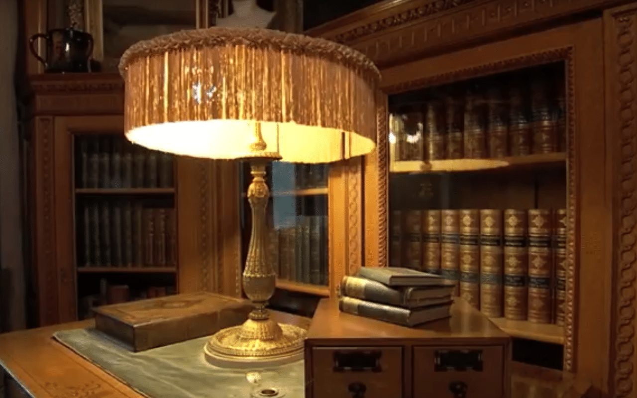 The Frick Building Pittsburgh video thumbnail sets the scene of a detailed antique styled glass enclosed bookcases set the backdrop for a intricate gold table lamp with a fringed shade set upon a wooden desk (video production by Merging Media).