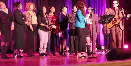 On stage, against a royal purple curtain, a casual choir sings behind a lead at the microphone accompanied by a acoustic guitar at the Mayor Peduto VIP Event Celebration (video production by Merging Media).