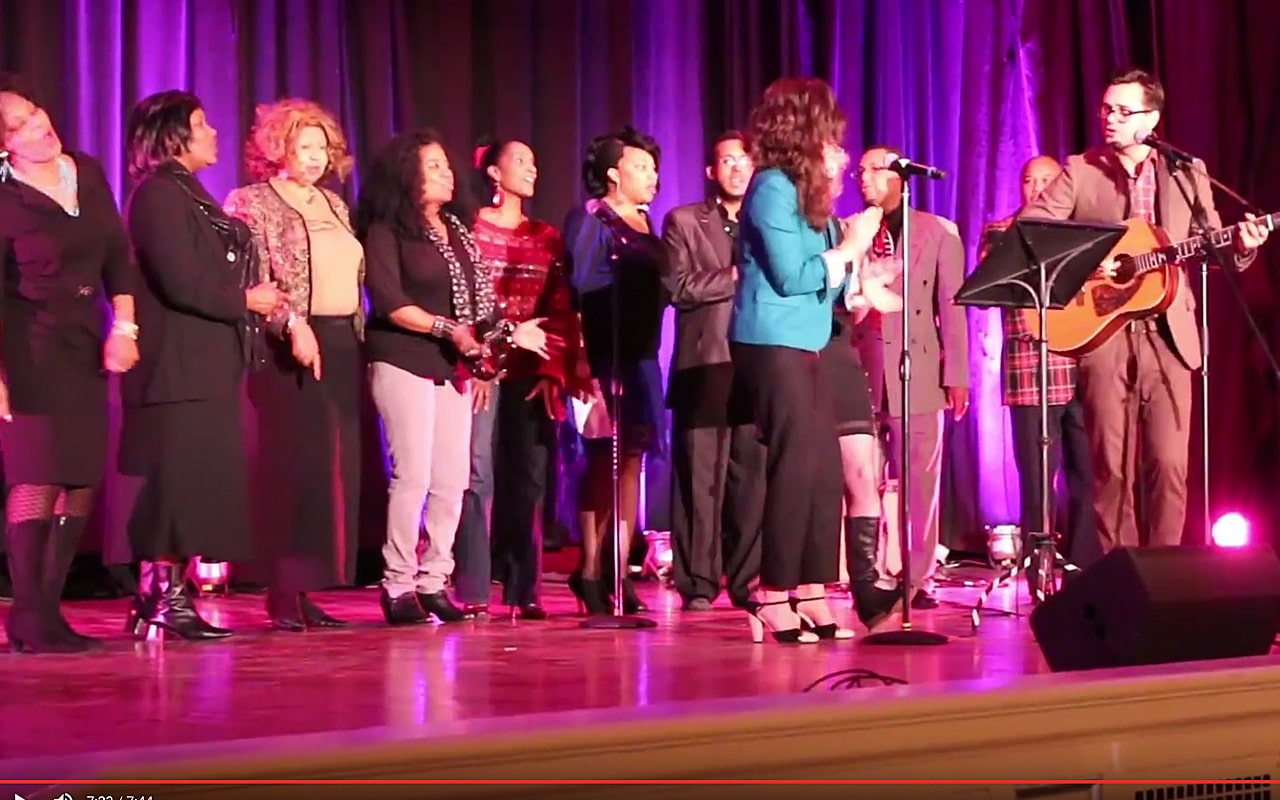 On stage, against a royal purple curtain, a casual choir sings behind a lead at the microphone accompanied by a acoustic guitar at the Mayor Peduto VIP Event Celebration (video production by Merging Media).