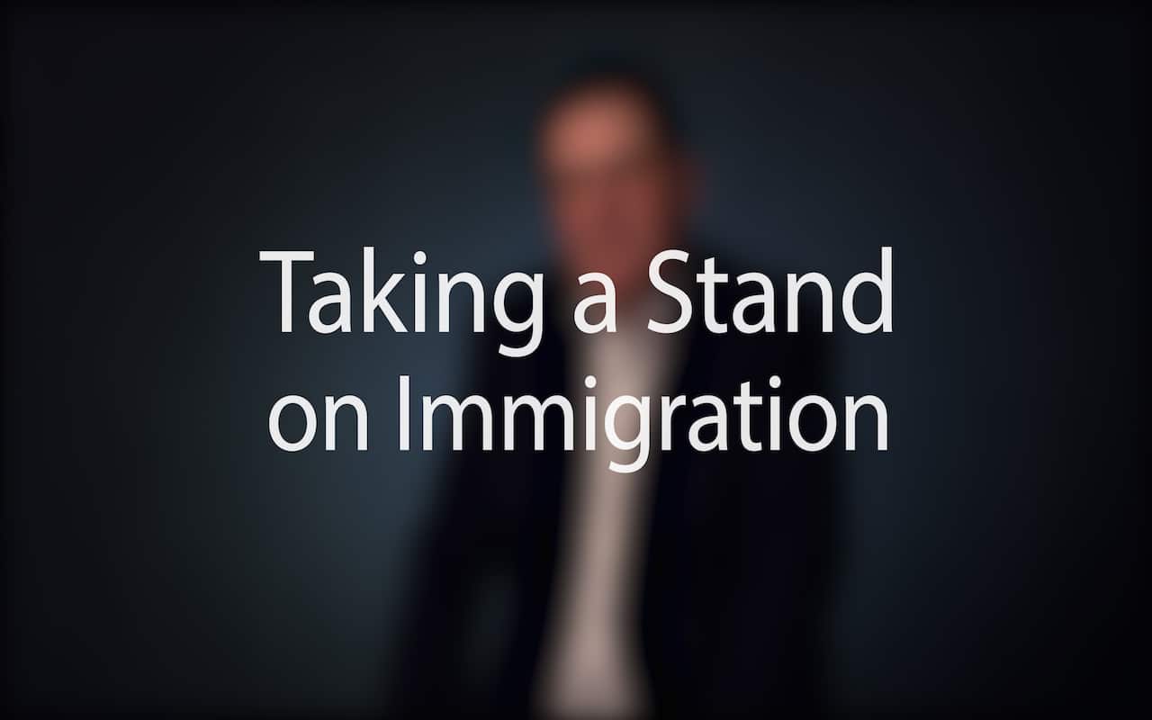 Mayor Peduto on Immigration campaign video; Blurred figure of a man in a suit sitting in front of a very dark blue background, the large word in the foreground in white text reads; Taking a Stand on Immigration (video produced by Merging Media).