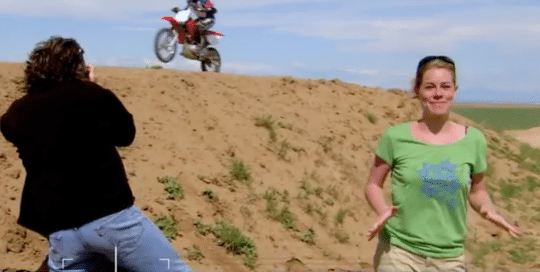 Host, Mara McFalls, with professional photographer, Jeff Swensen, uses the Panasonic Lumix digital camera to capture a photo of a fast moving motor bike racing across a sand dune in this video tutorial Panasonic Stopping The Action (video production by Merging Media).