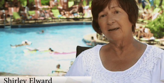 Perillo Tours Hawaii Testimonials video thumbnail features an older woman sitting for an interview about her Hawaii Perillo Tour in front of a backdrop of a busy hotel pool, title on the bottom left labels her as Shirley Elward from Ipswich, MA (video production by Merging Media).