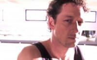 Bobrick Strategy Video thumbnail features a member of the mens rowing team wearing a black tank topped uniform, sitting in front of practice equipment and stares intently into the camera (video production by Merging Media).