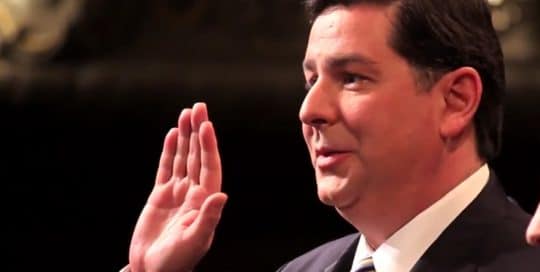 Mayor Peduto stands in his suit and tie at his inauguration with right hand in the air to be sorwn in as the mayor of Pittsburgh, Pennsylvania as recorded in Mayor Peduto Inauguration video (video production by Merging Media).