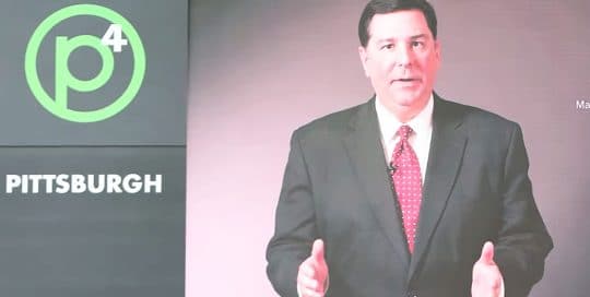 Mayor Peduto Fast Photo P4 Conference; Mayor Peduto stands in a black suit with a white shirt and red tie next to words that read P4 Pittsburgh (video production by Merging Media).