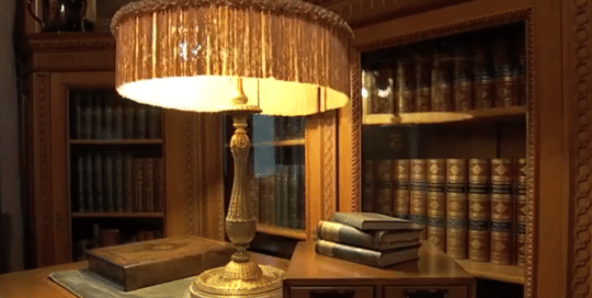 The Frick Building Pittsburgh video thumbnail sets the scene of a detailed antique styled glass enclosed bookcases set the backdrop for a intricate gold table lamp with a fringed shade set upon a wooden desk (video production by Merging Media).